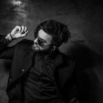 Black and white portrait of a fashionable man in sunglasses and suit posing confidently indoors.