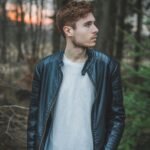 Young man in a black leather jacket standing in a forest during twilight, styled fashion portrait.